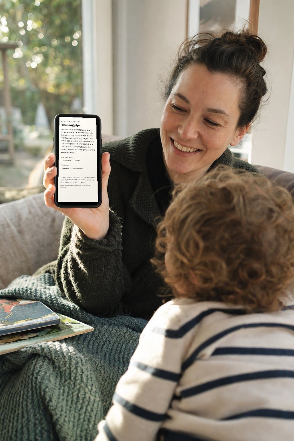 A parent at home smiling at a StoryLoop wall card on her phone, with a toddler leaning on her arm, seen from behind